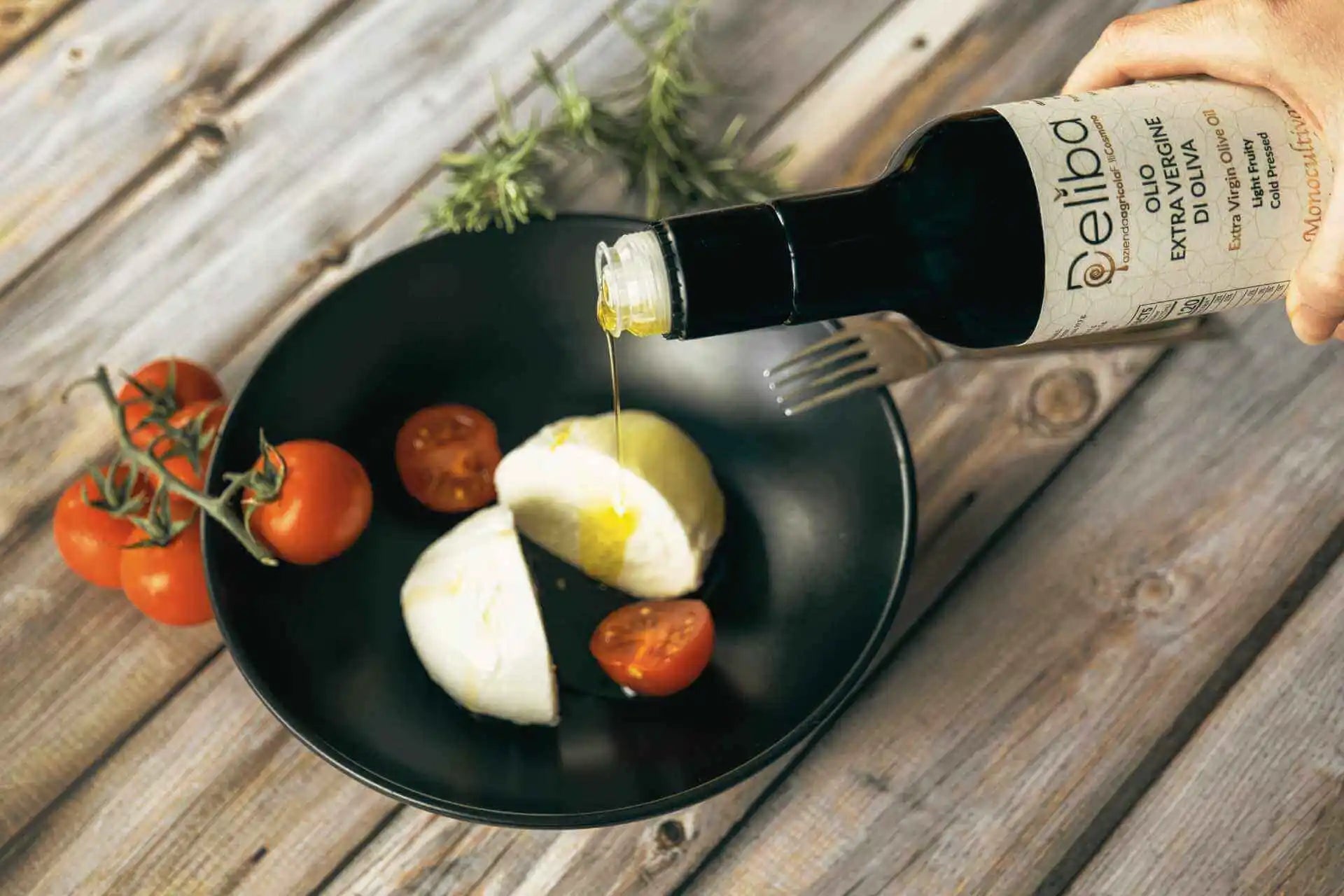 A person is pouring Deliba extra virgin olive oil on a halved buffalo mozzarella, accompanied by cherry tomatoes on a black plate, set on a wooden table. Some rosemary branches decorate the upper left corner.