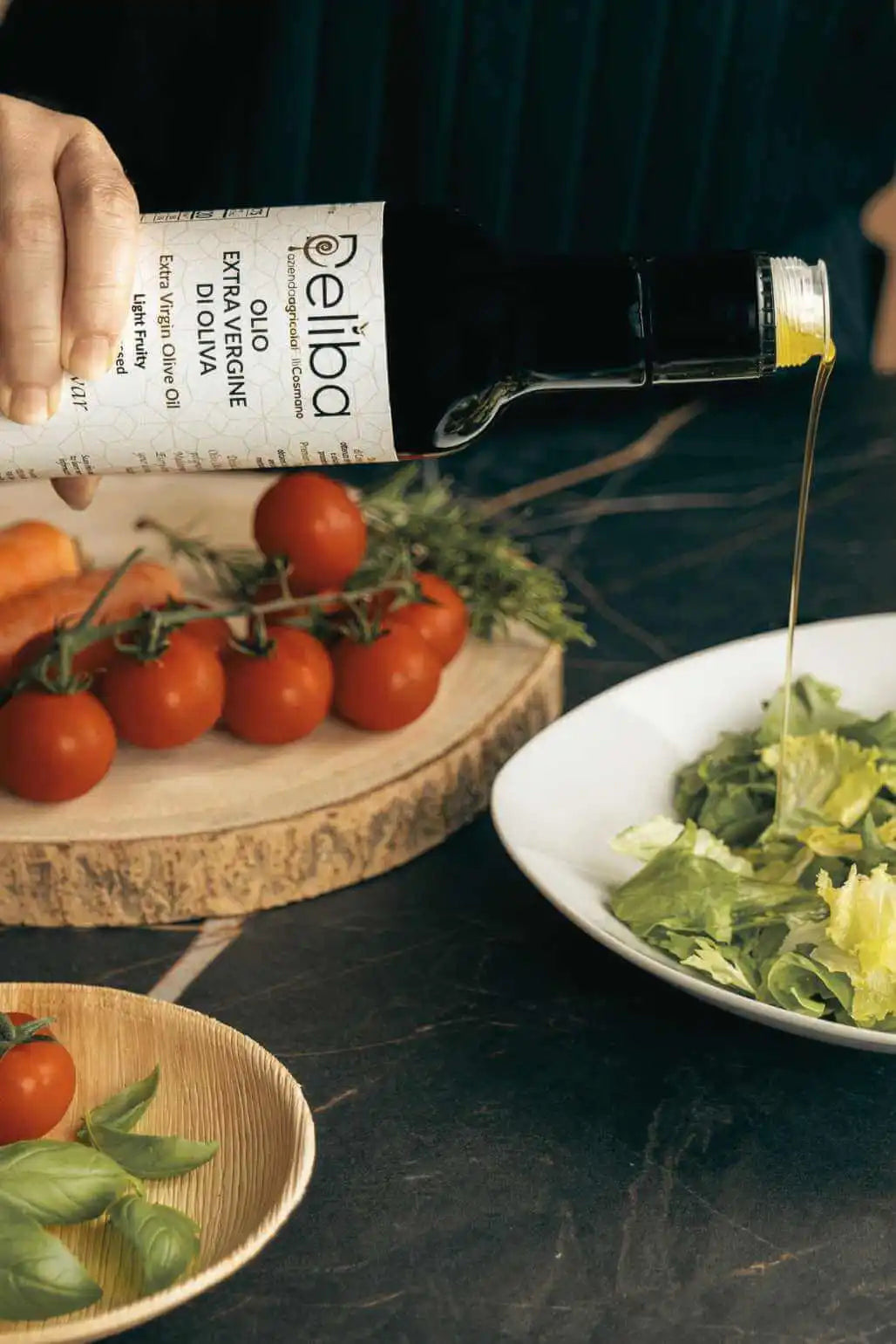 A hand is pouring Deliba olive oil from a bottle onto a plate of green salad. In the background, a wooden cutting board with cherry tomatoes on the vine and sprigs of rosemary, and a small dish holding fresh basil leaves, on a dark marble table.